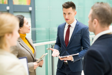 Group of modern business people discussing work standing in office building, focus on handsome young man talking to colleagues