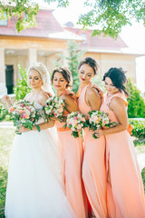 Stylish young smiling blonde bride with her bridesmaids in peach summer dress in the park on her happy wedding day. Women in the same color dress. All brunette one blonde.