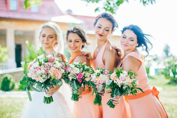 Stylish young smiling blonde bride with her bridesmaids in peach summer dress in the park on her happy wedding day. Women in the same color dress. All brunette one blonde.