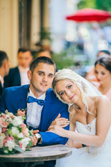 Happy fun newlyweds posing in street with bridesmaids & groomsmen. Bride with groom sitting at street cafe outdoor. 