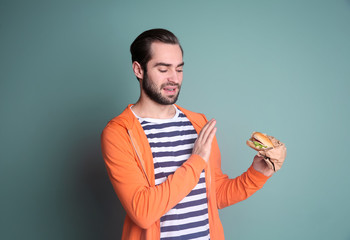 Handsome man refusing unhealthy burger on color background