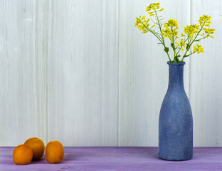A vase with a field of yellow flowers and apricots on the table.