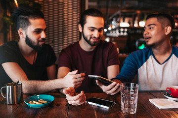 Group of mixed race young men using phone and talking in lounge bar. Middle-eastern friends having fun in cafe