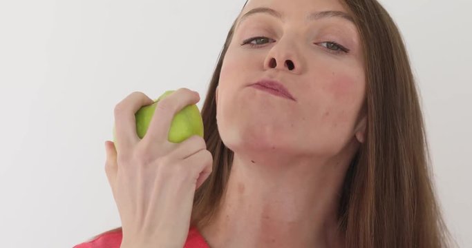 Young woman is eating a big green apple at white background. Healthy nutrition model eating fruit. Girl takes first bite and then offer bite to viewer and saying Wanna bite