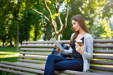 Obraz premium Motivated asian freelancer in the park. Girlon a bench, drinking coffee and working with tablet