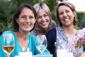 looking at the camera, three female friends in their 40s share a moment of complicity. They gathered around a table in the garden to share a meal with friends.