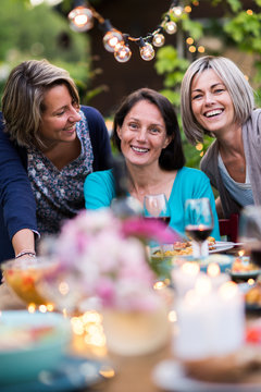 Summertime. Group Of Friends Gathered Around A Table In The Garden. Three Beautiful Women In Their 40s Pose For The Camera.