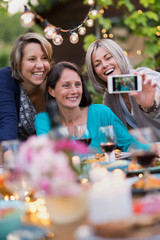 Summertime. Group of friends gathered around a table in the garden to share a meal. Three beautiful women in their forties taking a selfie