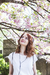 Young beautiful hipster redhead girl in white t-shirt and gray skirt listens a music from tapes in springtime blossom cherry trees garden. Retro tapes, spring time, go for a walk. 