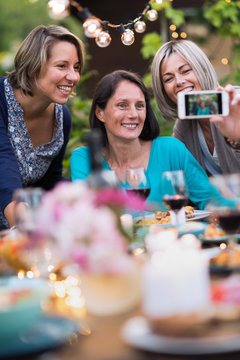 Summertime. Group of friends gathered around a table in the garden to share a meal. Three beautiful women in their forties taking a selfie