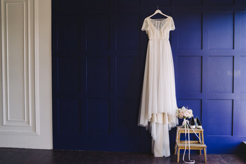 Wedding fashion. Bridal morning details. White lace dress hanging on blue wall, bouquet of flowers near it.