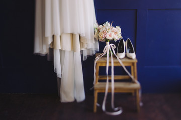 Wedding fashion. Bridal morning details. White lace dress hanging on blue wall, bouquet of flowers and leather shoes near it.