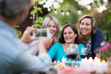 One summer evening friends gathered around a table in the garden for a good time. A man takes a picture of three female friends in their forties