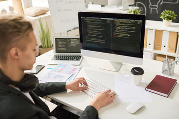 Portrait of contemporary young man coding at modern computer sitting at desk and working in startup project, focus on computer screen, copy space