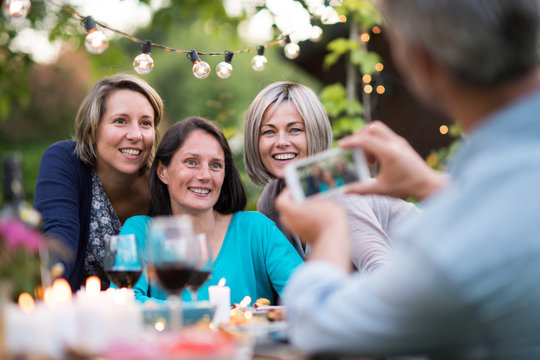 One summer evening friends gathered around a table in the garden for a good time. A man takes a picture of three female friends in their forties