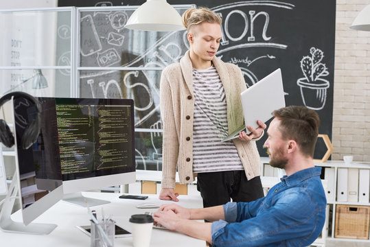 Portrait of creative young man standing by desk and holding laptop while writing code collaborating with colleague on startup project in modern office