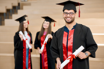 Happy smiling boy keeping university diploma. Graduate standing near young groupmates and looking...