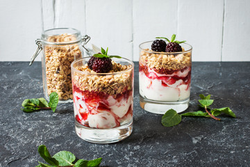 Healthy layered dessert with yogurt, granola, jam, blackberry in glass on stone background.