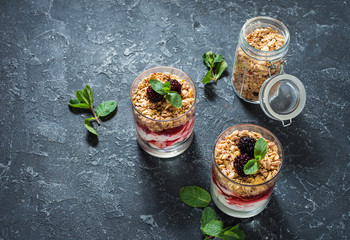 Healthy layered dessert with yogurt, granola, jam, blackberry in glass on stone background