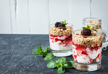 Healthy layered dessert with yogurt, granola, jam, blackberry in glass on stone background.