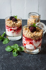Healthy layered dessert with yogurt, granola, jam, blackberry in glass on stone background.