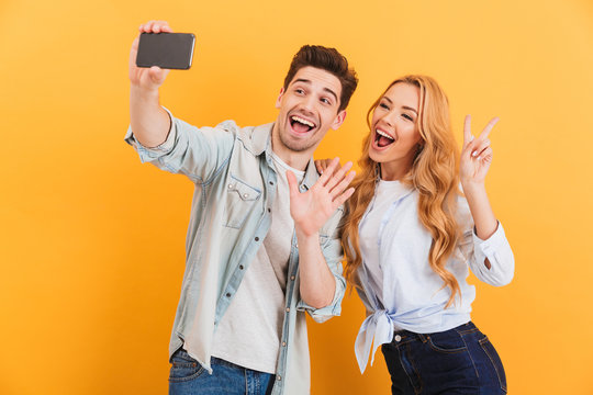 Portrait of happy man and woman taking selfie photo on cell phone while gesturing at camera with fingers, isolated over yellow background