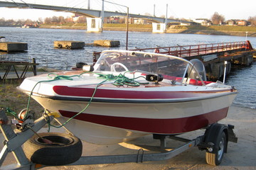 Descent boats on the water – red and white boat with an outboard motor on the trailer near the water fall at the background of the river, Wharf and bridge © Ilya