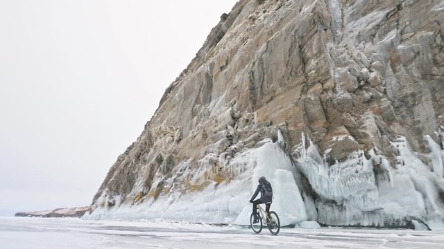 Man is riding bicycle near the ice grotto. The rock with ice caves and icicles is very beautiful. The cyclist is dressed in gray down jacket, cycling backpack and helmet. The tires on covered with