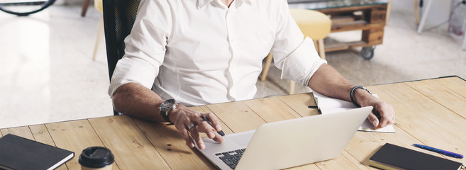 Adult bearded man working on mobile laptop computer while sitting at wooden table.Cropped. Wide.