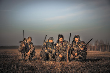 Men hunters group team portrait in rural field posing together against sunrise sky during hunting...