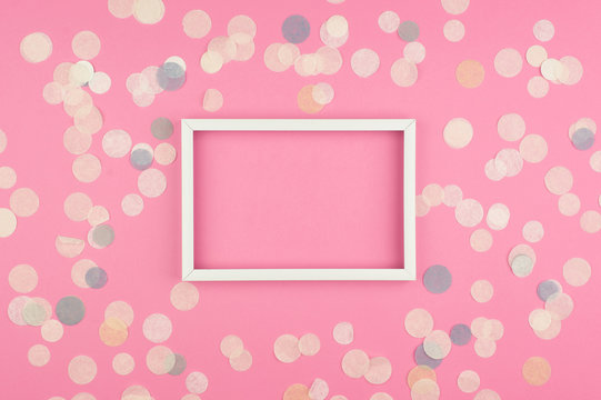 White picture frame and confetti on pink background. Top view, flat lay. Mockup for party or birthday invitation.