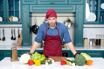 Cook stand at kitchen table. Man in chef hat and apron in kitchen. Vegetables and tools ready for cooking dishes. Vegetarian menu and healthy diet. Food preparation and cooking recipes