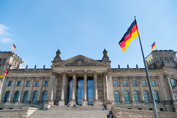 Obraz premium Niemieckie flagi powiewające na wietrze w słynnym budynku Reichstagu, siedzibie niemieckiego parlamentu (Deutscher Bundestag), w słoneczny dzień z błękitnym niebem i chmurami, centralna dzielnica Berlin Mitte, Niemcy