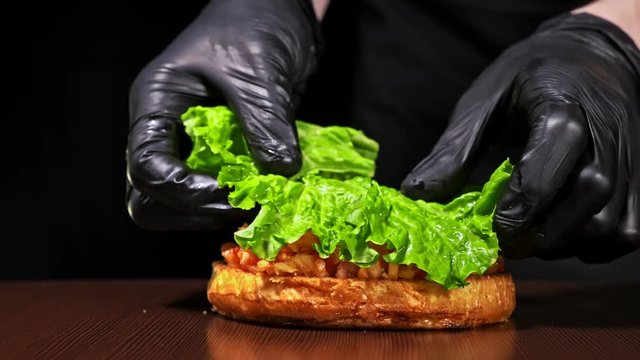 Burger is cooking on black background in black food gloves. Very luscious air bun and marbled beef. Restaurant where each burgers is cooked by hand. Put salad lettuce and pickle cucumber on bun. Not
