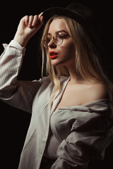Fashionable blonde girl in glasses and hat, wearing shirt with naked shoulders, posing with dramatic light at studio