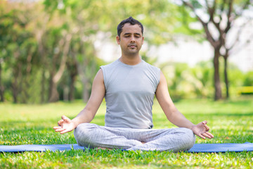 Serene Indian guy practicing yoga outdoors. Young yoga practitioner working out and sitting in...
