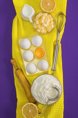 Baking flat lay composition with white eggs, yolk, a cup of flour, a   cup of butter, milk, lemon and kitchen tool sieve, rolling pin and   whisk on yellow and violet background.
