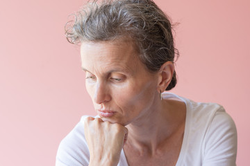 Head and shoulders view of middle aged woman with head on hand looking down against pink background (selective focus)