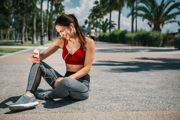 Cheerful female athlete is making selfie on smartphone. She is sitting on the road in summer park and laughing. Girl is wearing earphones