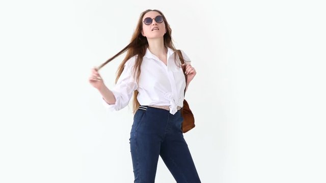 fashion of modern youth. stylish girl posing against white wall in jeans, white shirt, with leather backpack and glasses.