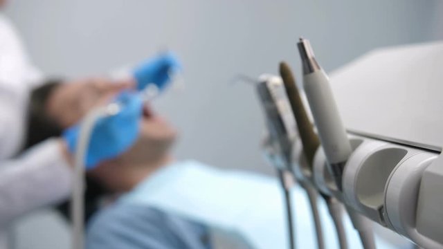 Close-up female dentist's hand in gloves taking dental drill machine on the foreground and starts treating male patient's teeth blurry on the background. Dental treatment at clinic
