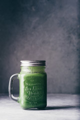 Green smoothie in jar with apple,baby spinach, parsley and banana, over a green round plate on a gray board against a gray background with parlsey leaves.