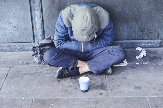 Homeless Man Sitting On The Street In The Shadow Of The Building And Begging For Help And Money. Problems Of Big Modern Cities.