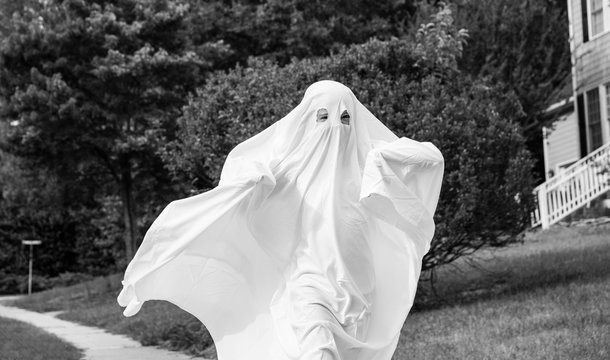 A black and white photo of a child in a ghost costume made out of a bedsheet running down the sidewalk.