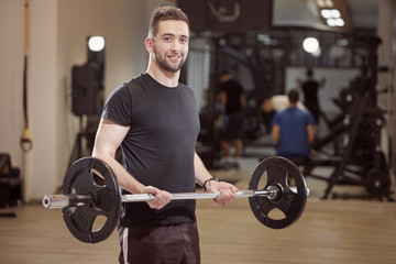 Fototapeta na wymiar one young man posing, ordinary, average looking, holding barbell with weights, exercise in gym. Unrecognizable people behind (out of focus).