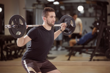 Fototapeta na wymiar upper body shot, squat exercise, young man holding barbell with weights on back shoulders. Unrecognizable people behind in gym (out of focus)