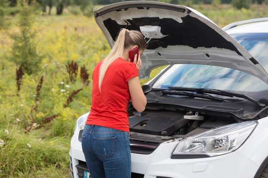 Rear view image of young woman looking under the hood of broken car and calling service
