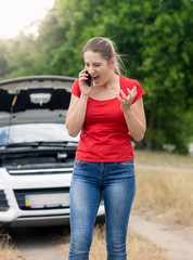 Angry young woman standing at broken car in field and shouting in mobile phone