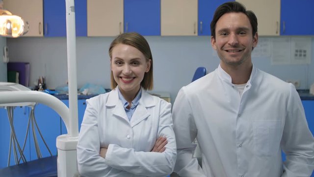 Two medical staff coworkers in white lab coats at doctor's office looking at camera and smiling with radiant toothy smiles. Handsome young male doctor and beautiful female dentist smiling