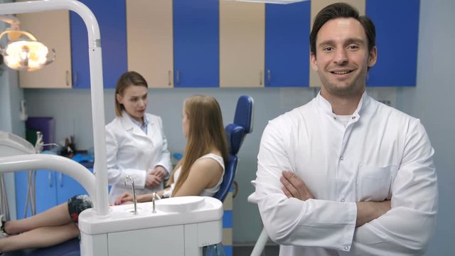 Handsome young adult male dentist standing and smiling to camera with arms crossed in lab coat. Dentist's female assistant talking to patient sitting in dental chair at the background at dental clinic